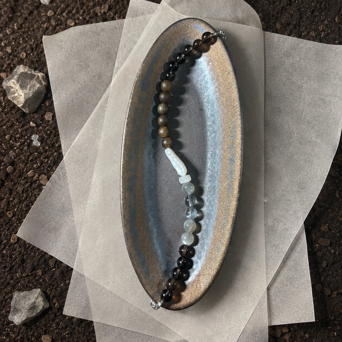 Strata Pale Drift Bracelet styled on a dark surface with pearl, moonstone, smoky quartz, and agarwood details